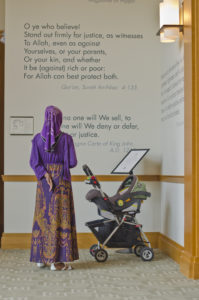 A person wearing a hijab stands next to a baby carriage in front of a wall with quotes. 