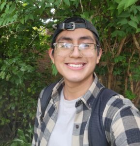 A person smiles at the camera with greenery in the background. He is wearing a backwards hat and flannel. 