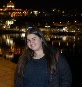 A person stands smiling in front of a city background at night. She has long dark hair and is wearing a black shirt.